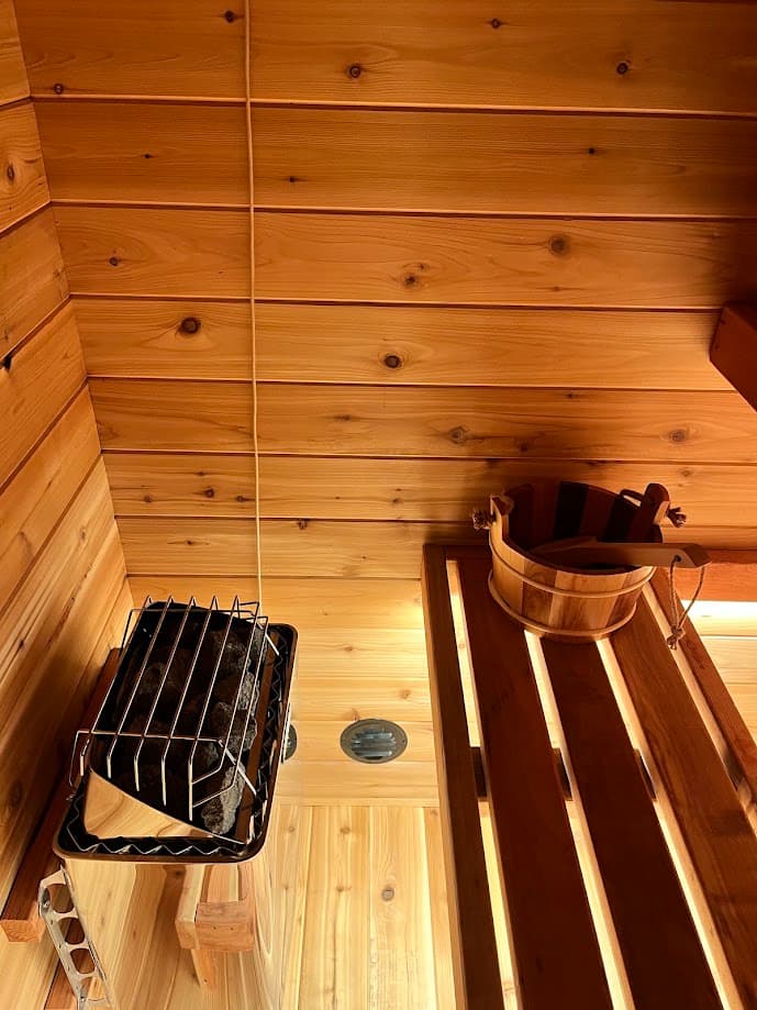 A round intake vent installed low on the sauna wall right beside the heater, so incoming air is warmed by the stove as it enters