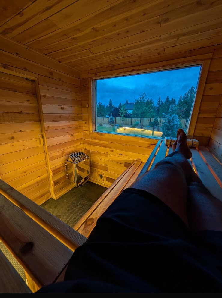 View from top bench at dusk – feet up looking through picture window at trees and twilight sky, Harvia heater and cedar benches below